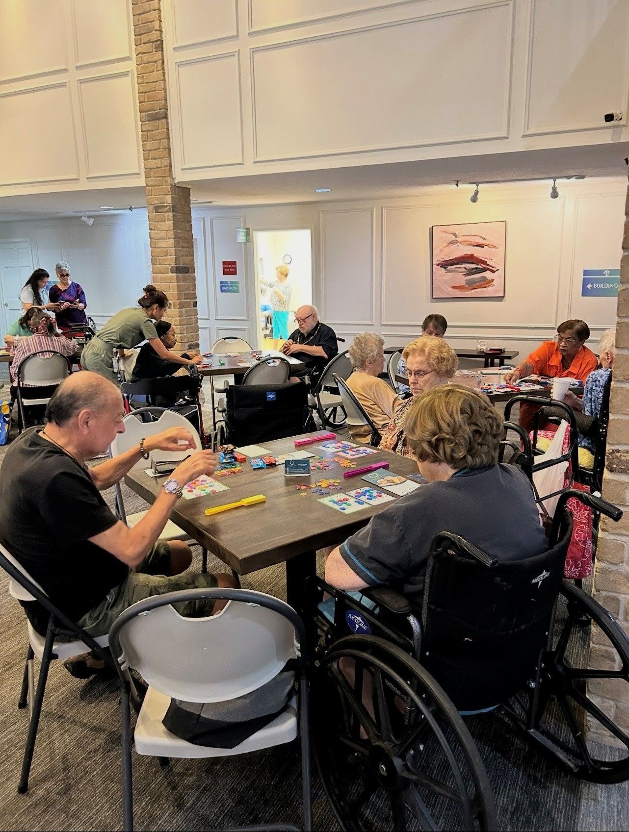 People sit at tables in a bright, modern room, engaging in tabletop activities and games with assistance from staff.