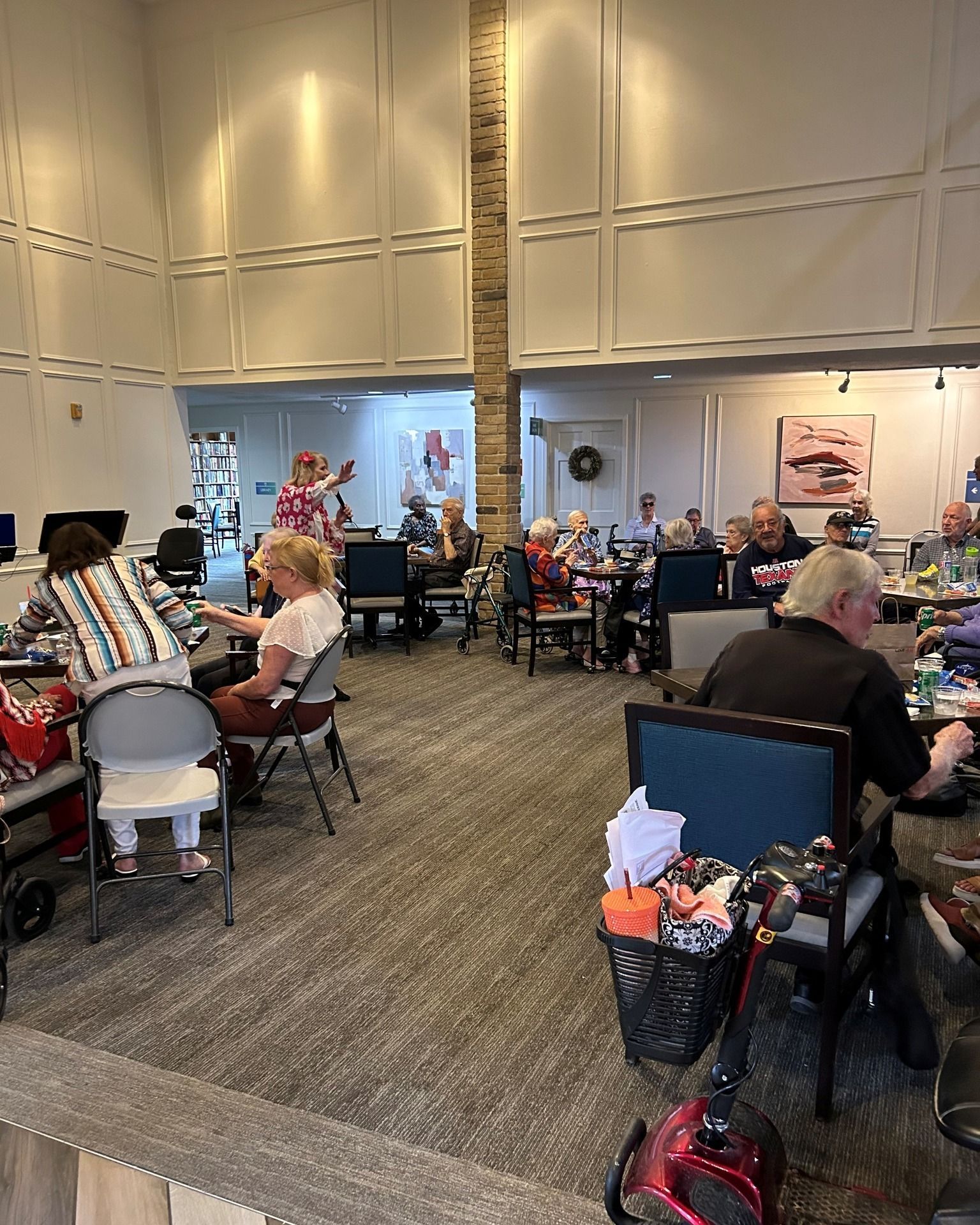 People seated at tables in a room, some with mobility aids. A person waves. Beige carpet, light walls.