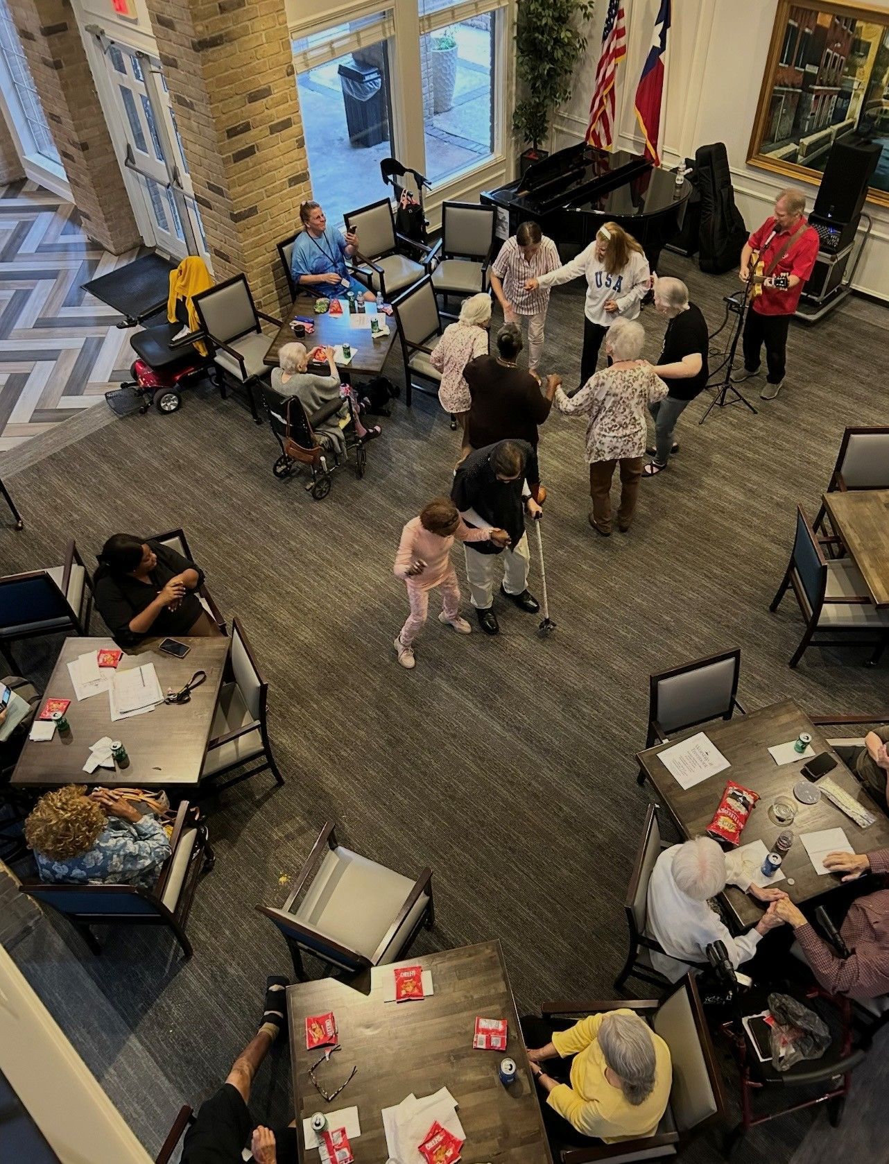 A group of people dance in a community room while a man plays guitar, with others seated at tables nearby.