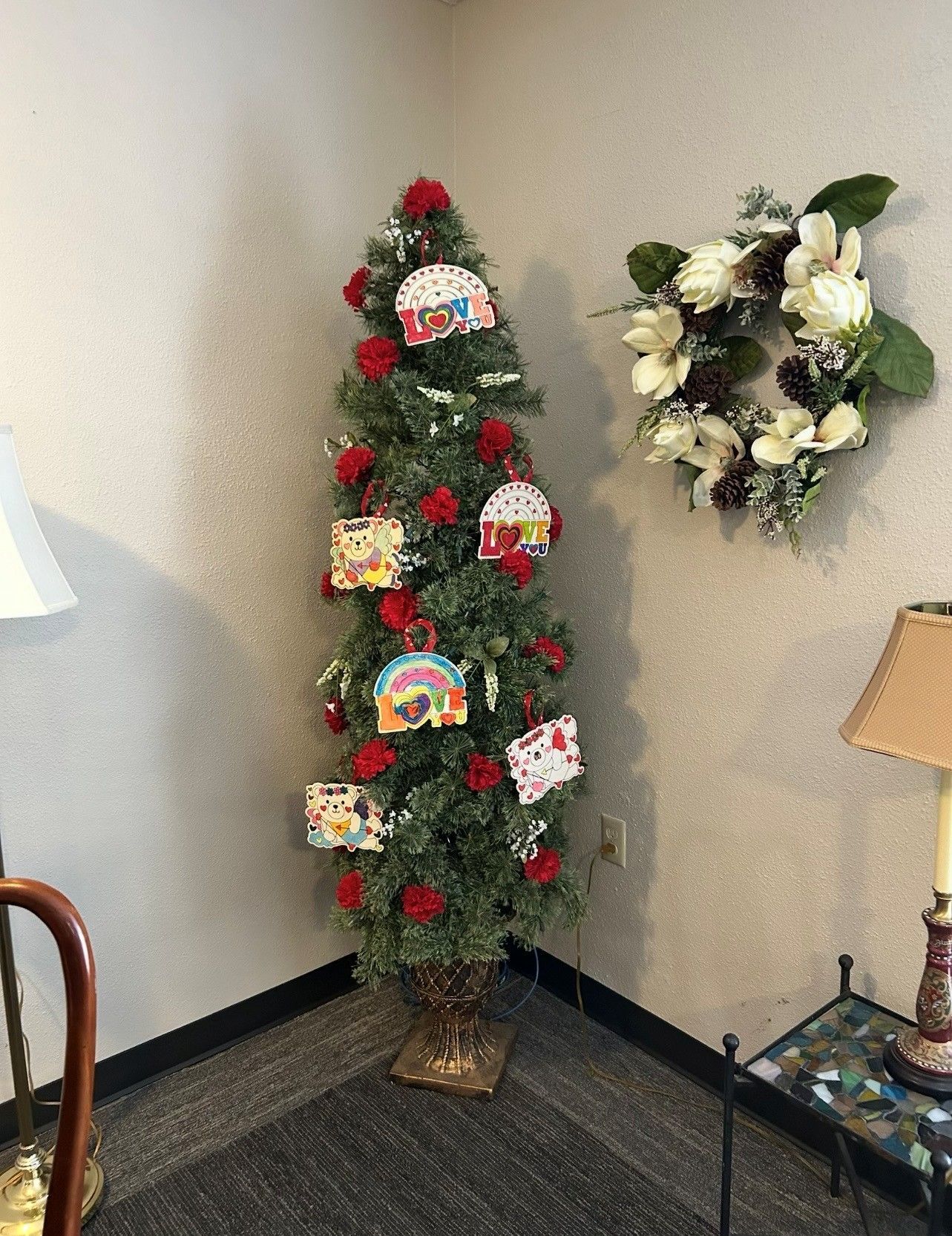 A tall, thin green tree decorated with red roses and colorful ornaments in a corner, with a flower wreath on the wall.