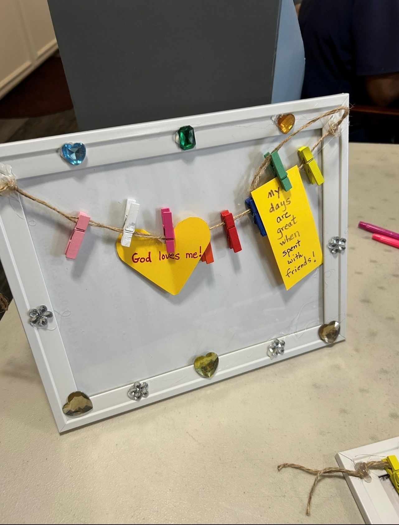 A white frame with a string across it holds yellow paper notes decorated with colorful clothespins and gem embellishments.