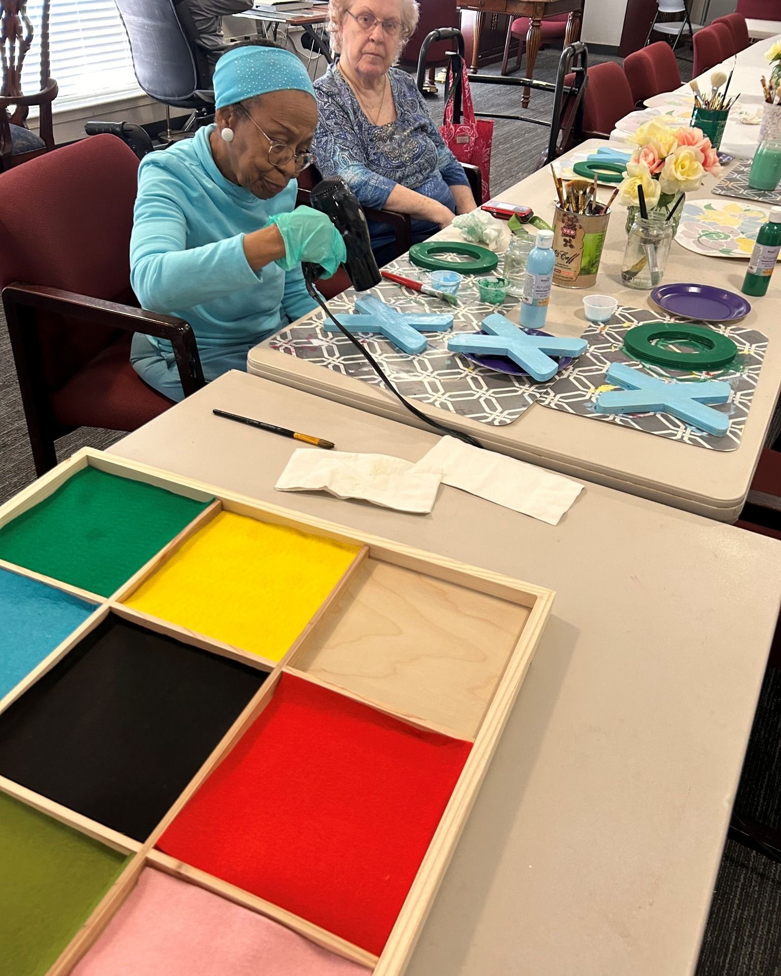 Two people at a table painting crafts. Woman in blue cap uses a heat gun, with colorful crafts and supplies.