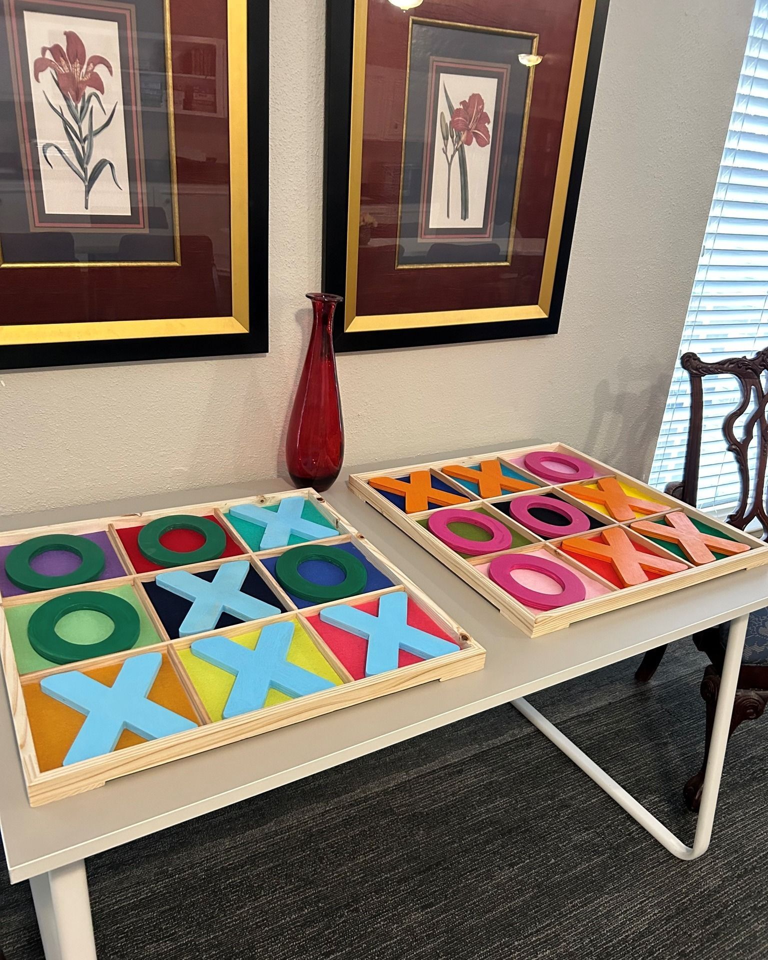 Two colorful tic-tac-toe games on a table, with framed art and a vase in the background.