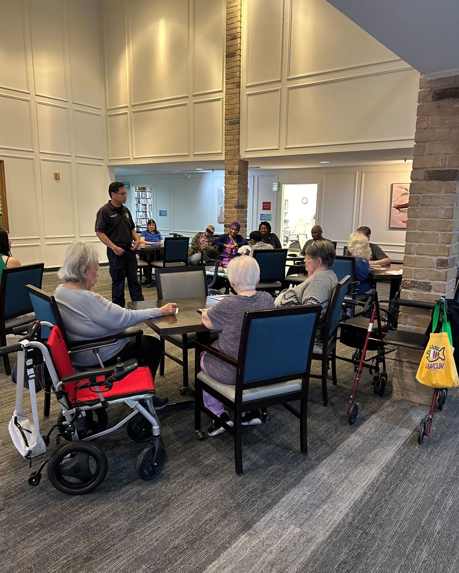 A man speaks to a group of seniors seated at tables in a large room. Some use wheelchairs or walkers.