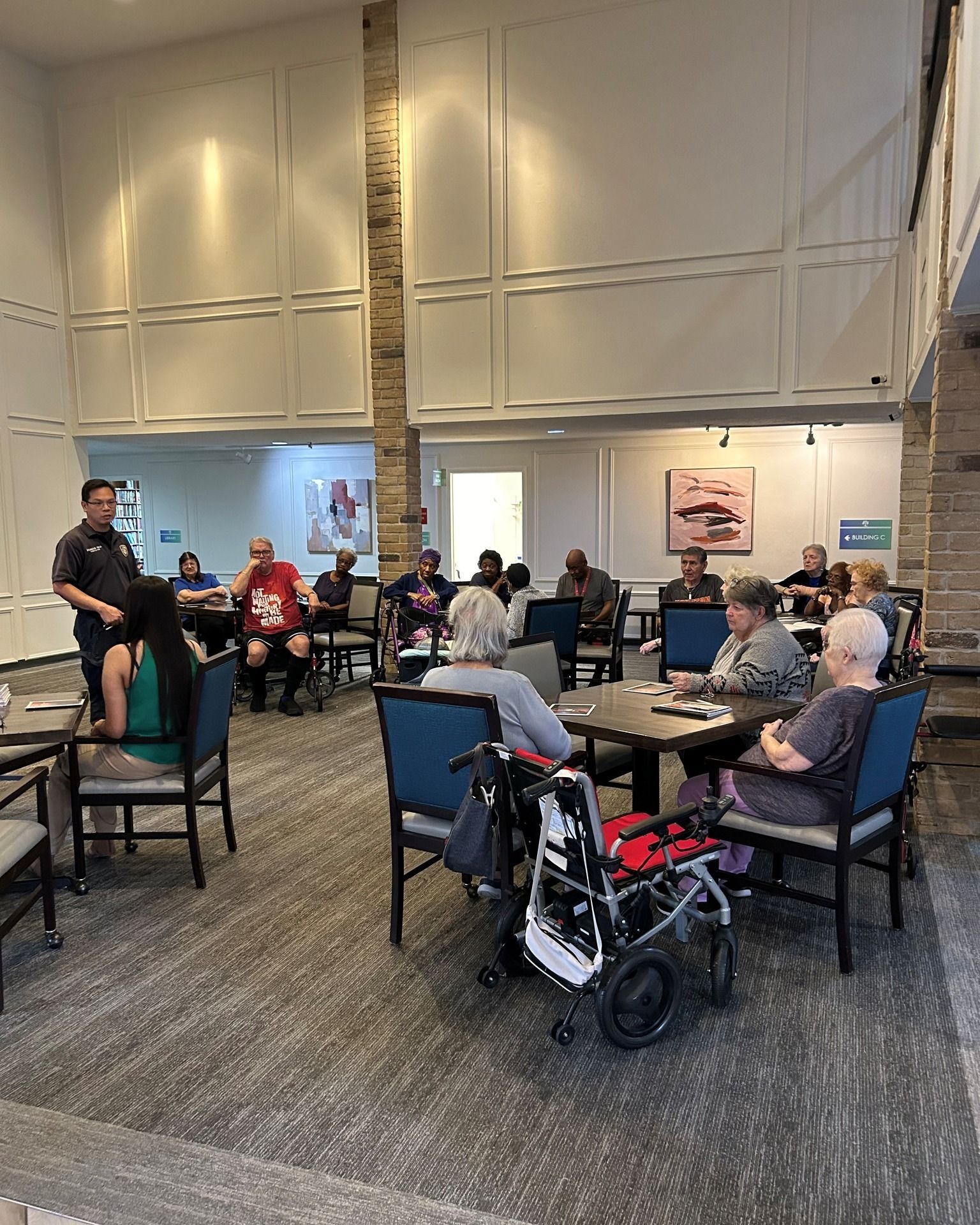 People seated around tables in a room, some in wheelchairs. A person stands and speaks to the group.
