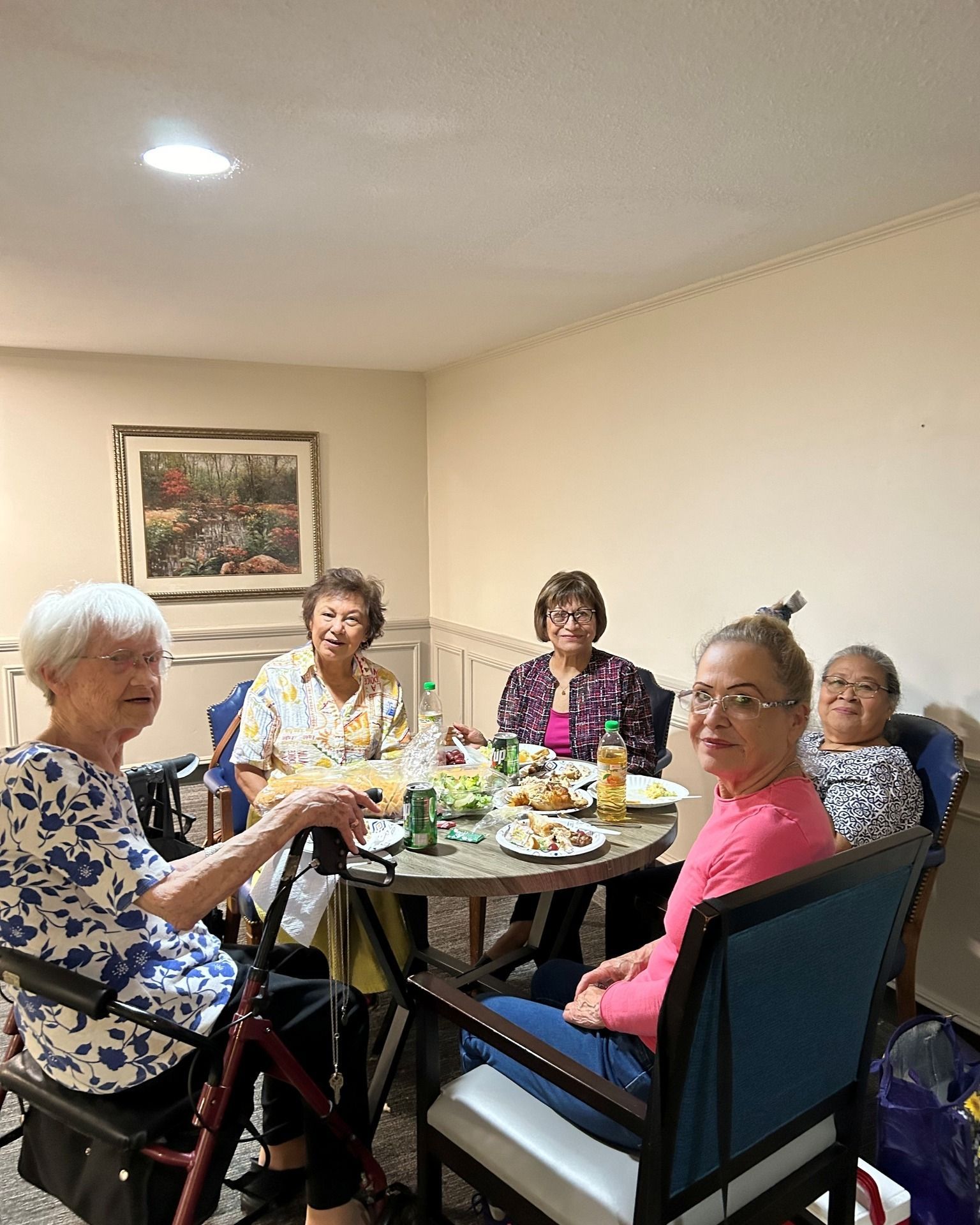 Five people seated around a table, enjoying a meal indoors. Light-colored walls and a painting are visible.