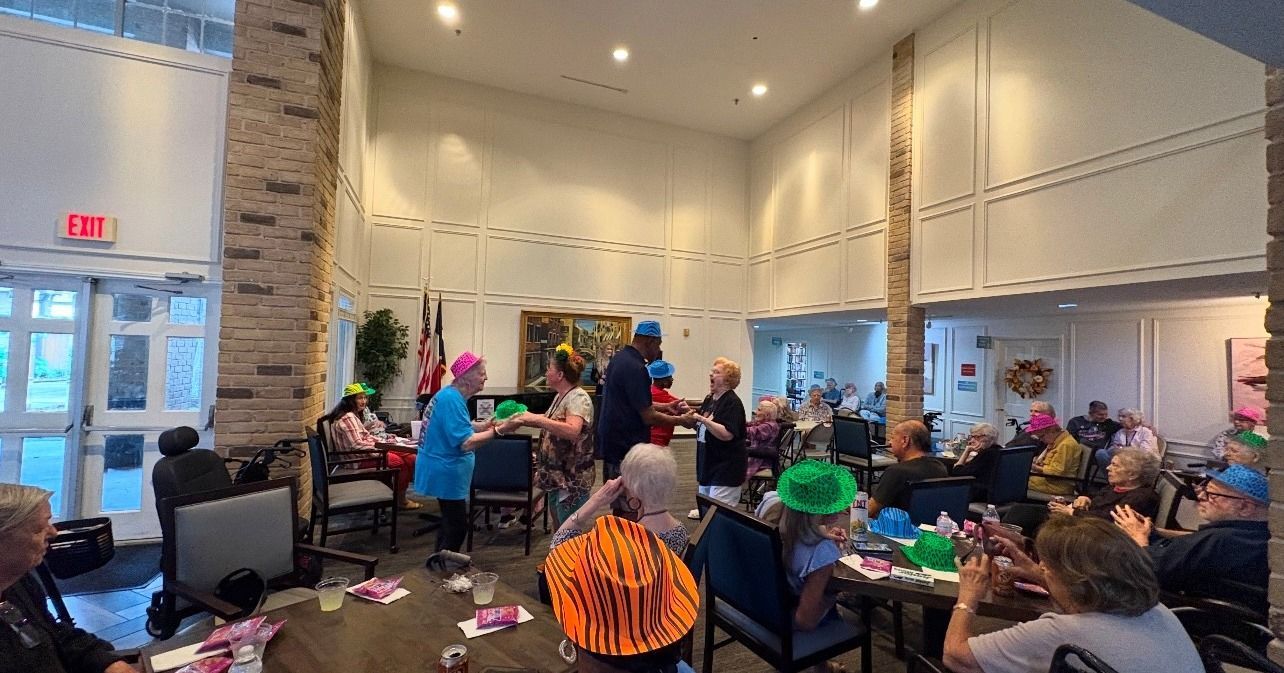 People with party hats in a room, celebrating. A person in a suit is in the center. Some are seated, some are standing.
