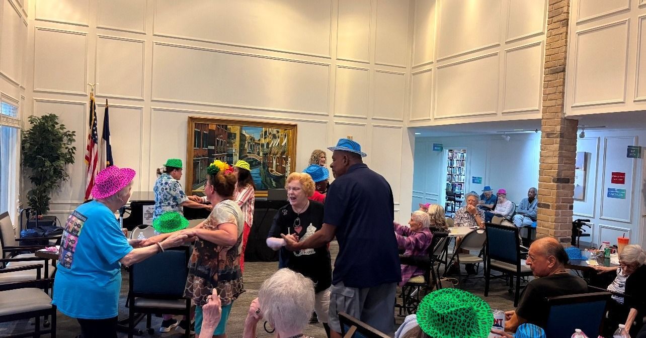 Group of people dancing in a room, some wearing colorful hats. Others seated at tables.