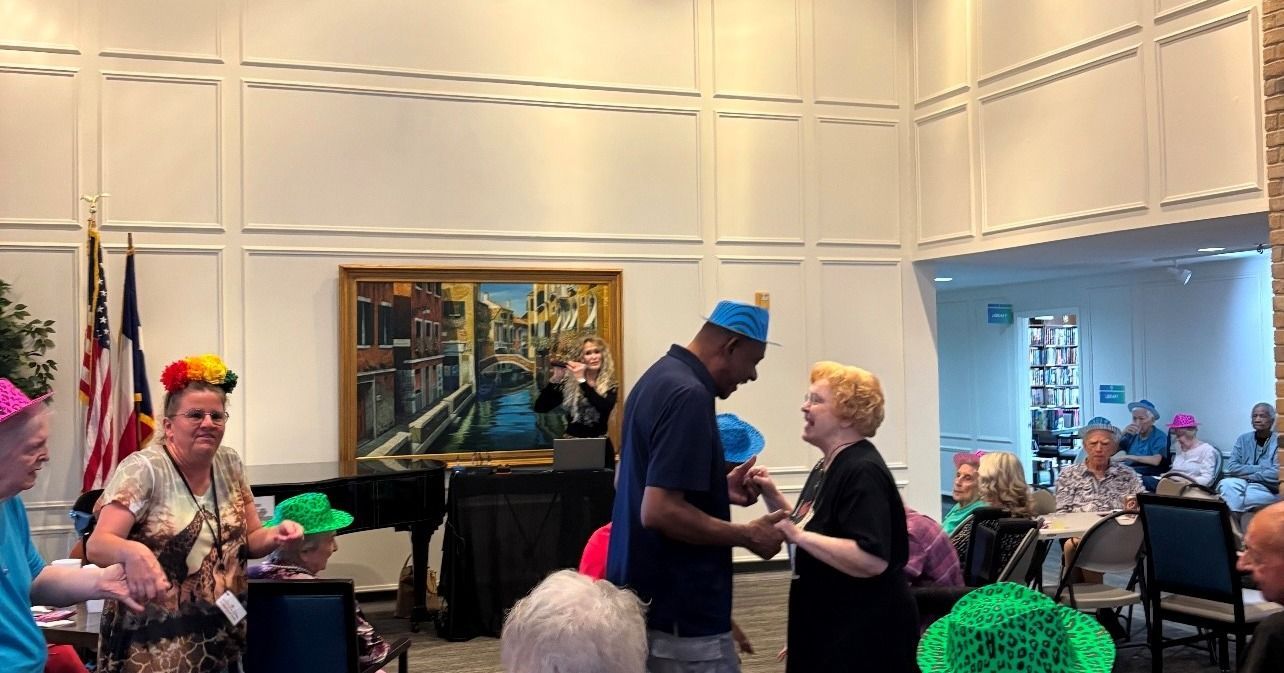 People in a room, some wearing colorful hats, a man and a woman shaking hands, celebrating.