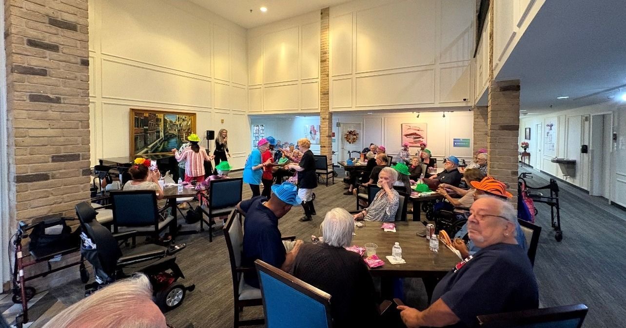 Seniors in a brightly lit room gather, some wearing hats, seated at tables, likely enjoying an event.