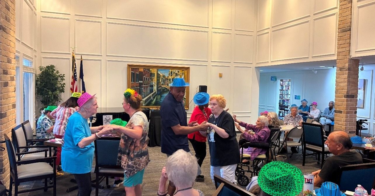 Seniors dancing in a hall. Some wear colorful head coverings. The room has tables and decorations.