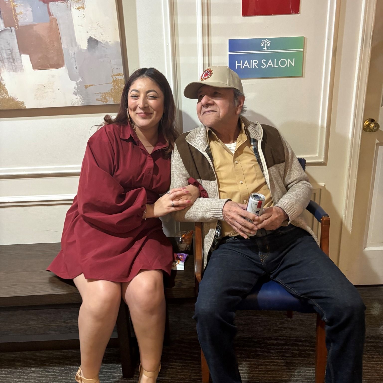 Woman in burgundy dress and man in a hat sit together indoors. Hair salon sign visible.
