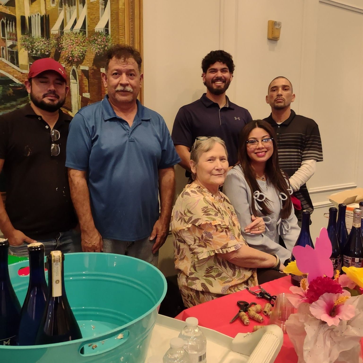 Group of people smiling, standing behind a table with wine bottles and decorations in a well-lit room.