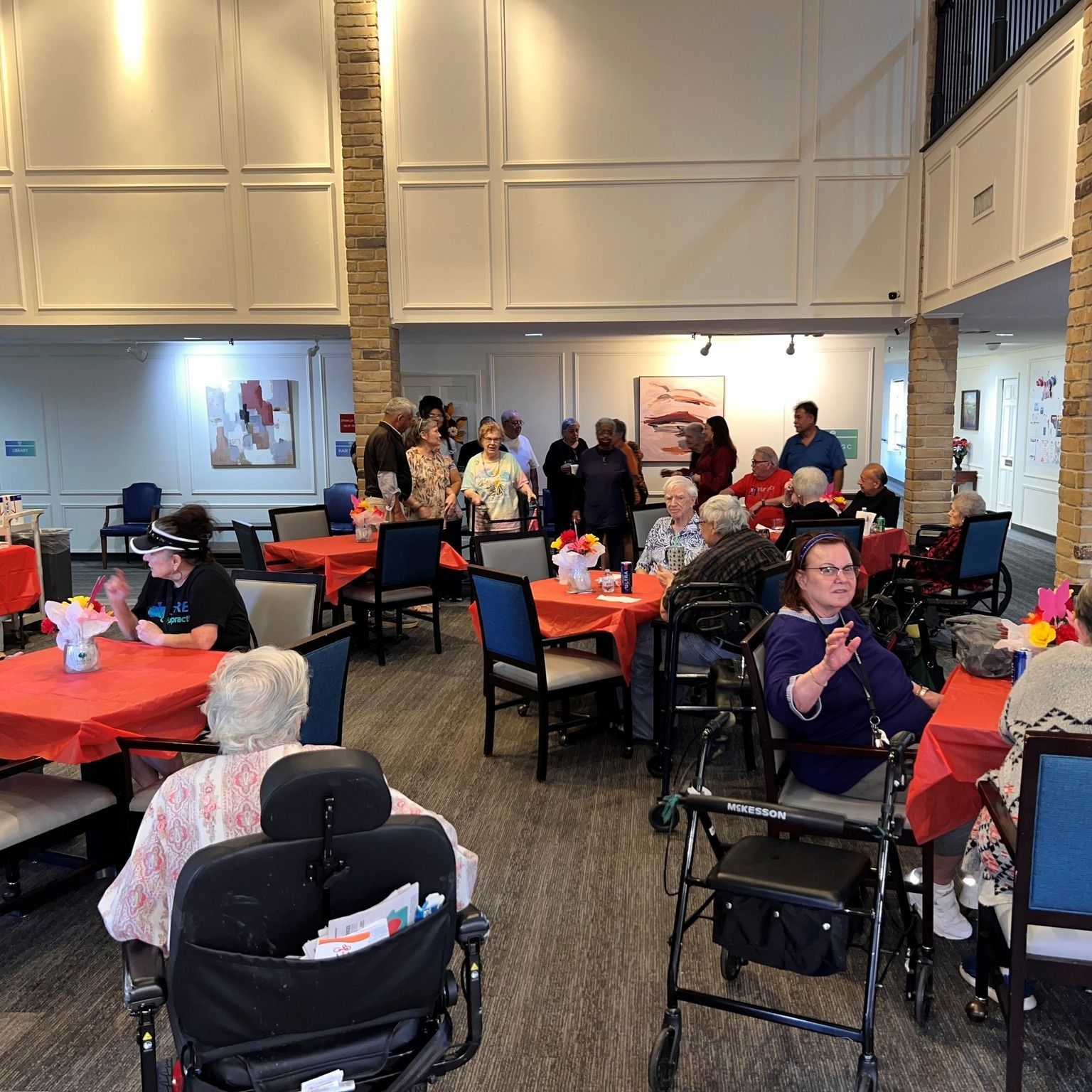 Seniors at tables in a brightly lit room with red tablecloths, some in wheelchairs, appear to be socializing.