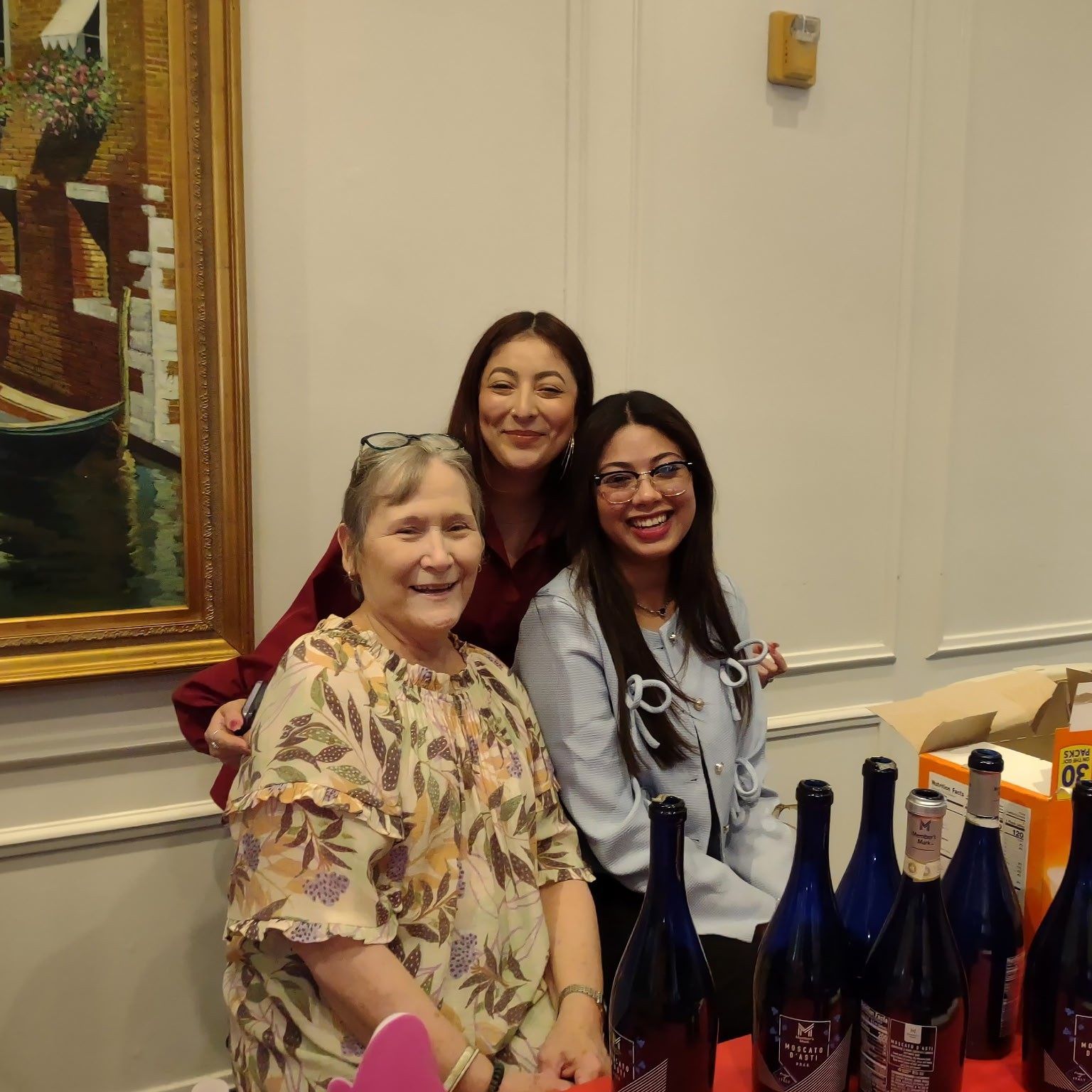 Three women pose at a table with wine bottles. One smiles, two are behind. Art on wall.