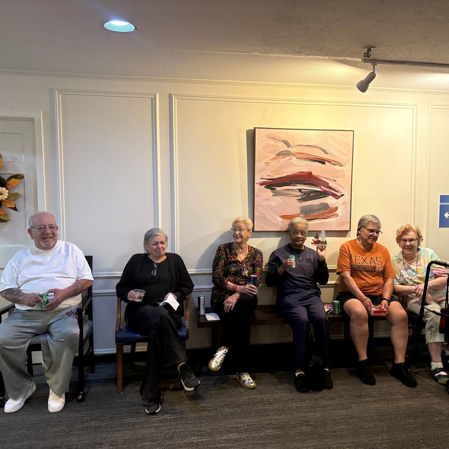 A group of older adults sit in a waiting area. Some smile, holding drinks. There's artwork on the wall.