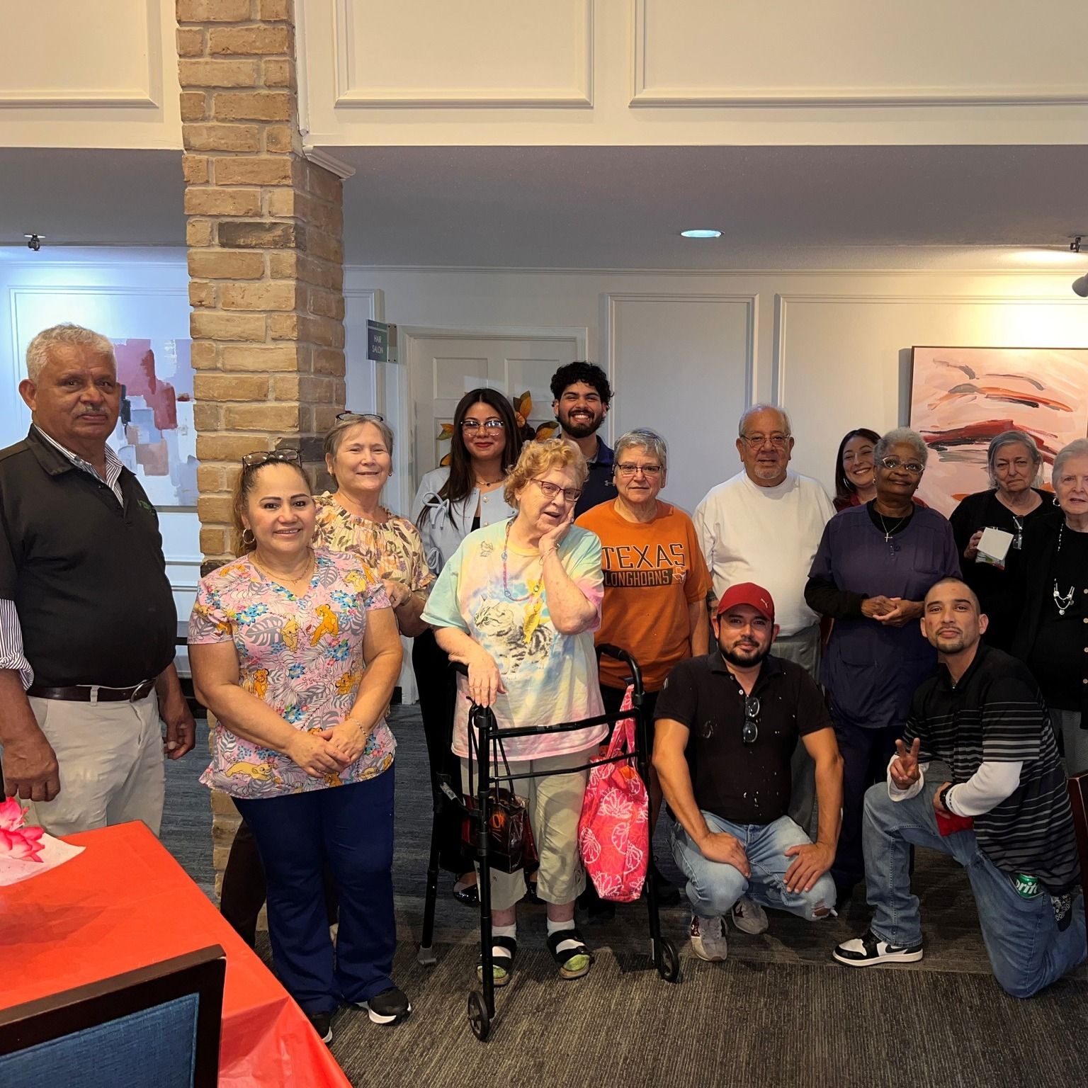 Group of people smiling, posing for a photo indoors. Some are elderly, one uses a walker.