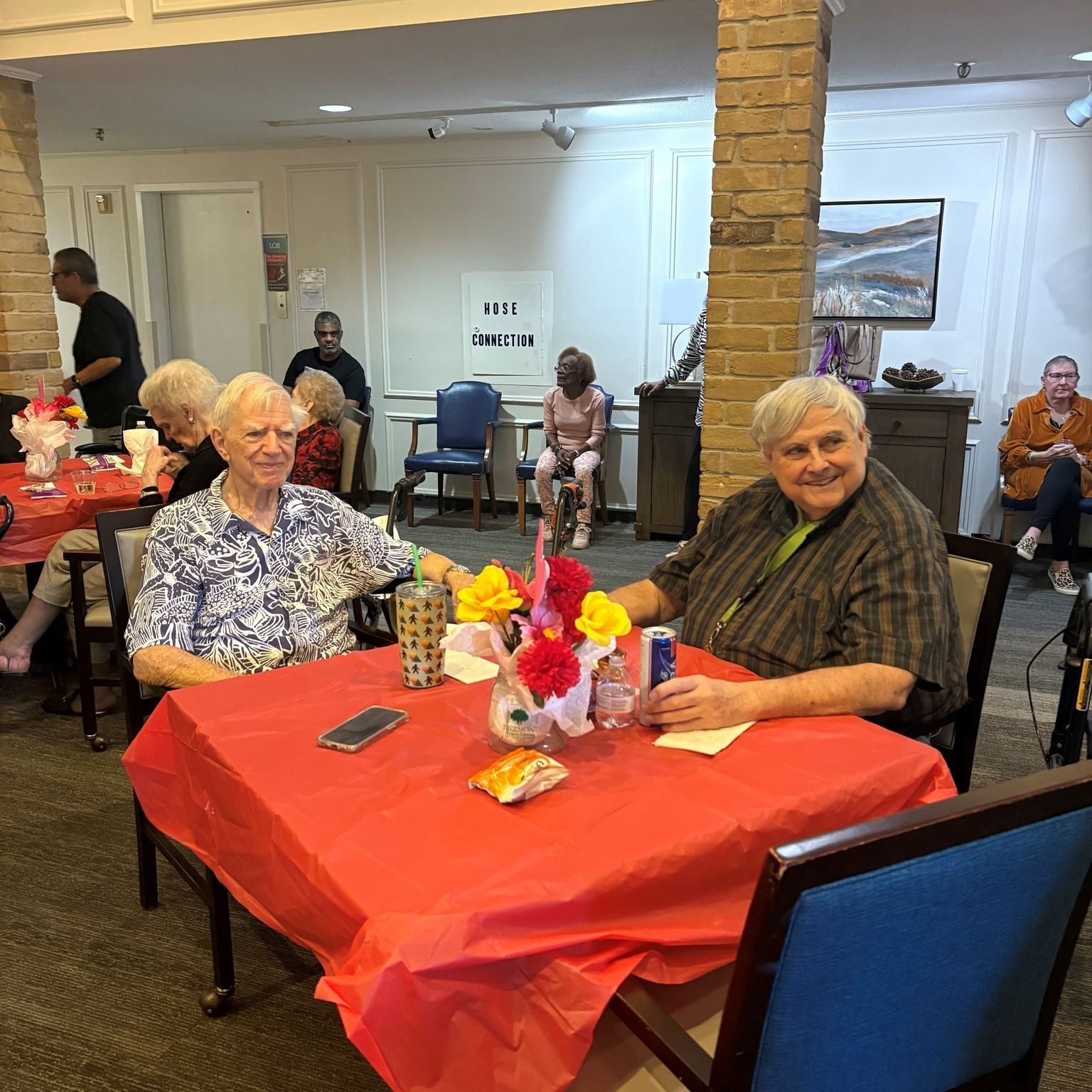 People at tables in a room, two men smiling at a table with a red cover, flowers, food and drinks.