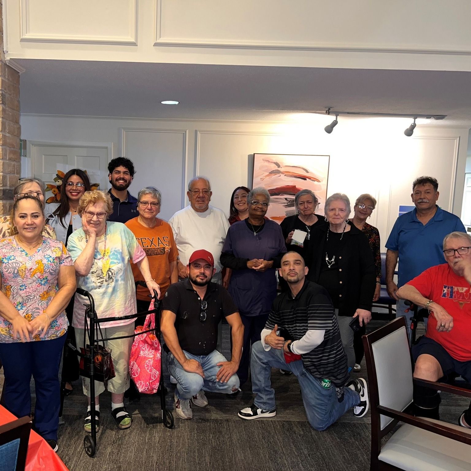 Group of people indoors, some smiling, posing for a photo. A few have mobility aids. Well-lit room.
