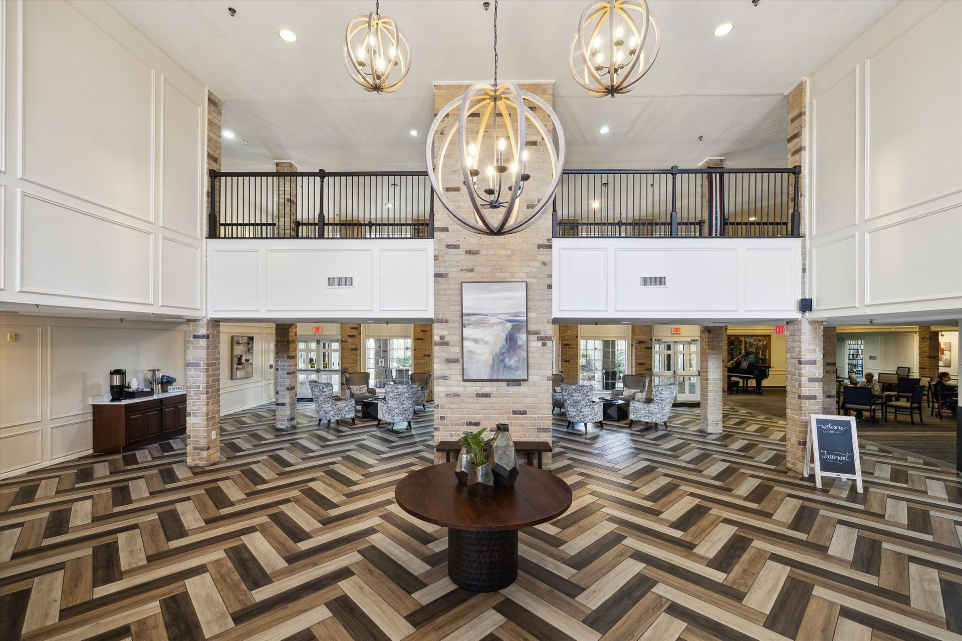 Lobby with herringbone floors, two-story ceiling, decorative light fixtures, seating areas, and a second-floor balcony.