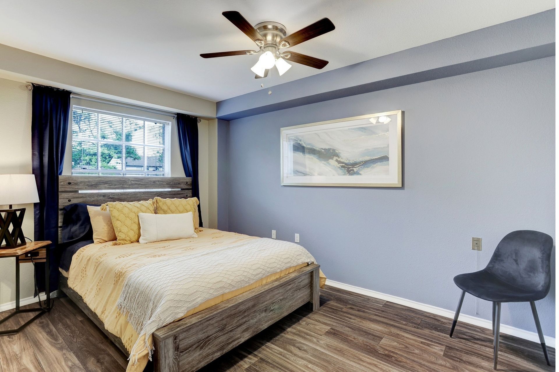 Bedroom with bed, blue wall, window, dark blue curtains, art, chair, and ceiling fan.