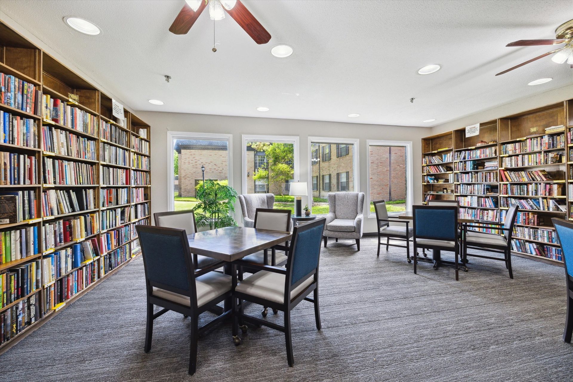Library with bookshelves, tables, chairs, and windows overlooking a courtyard.