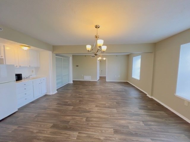Empty apartment interior with kitchen, dining, and hallway. Brown wood-look flooring, white cabinets, and neutral walls.