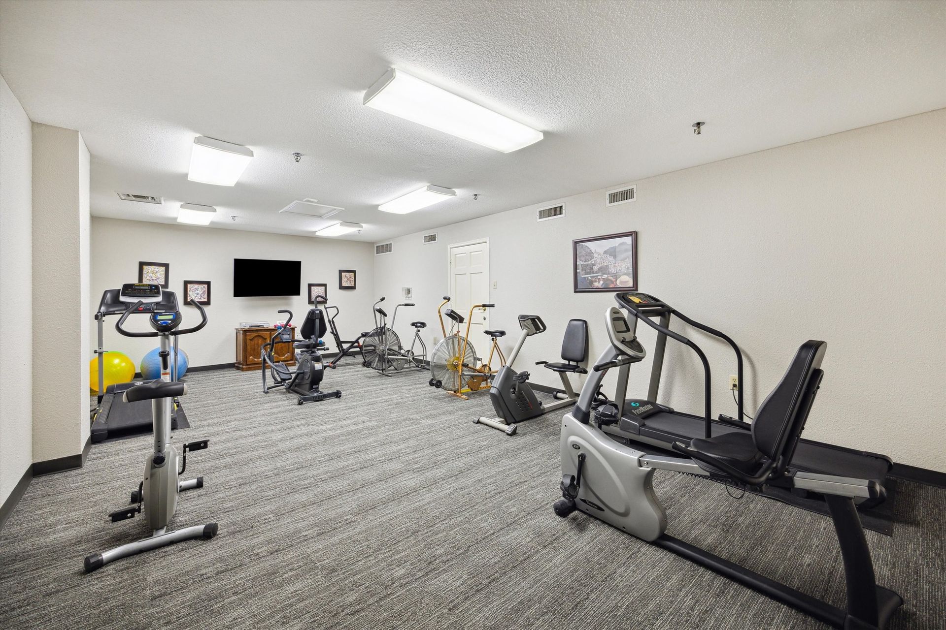 Gym with exercise equipment, including bikes and treadmills, on a carpeted floor. A TV is on the wall.