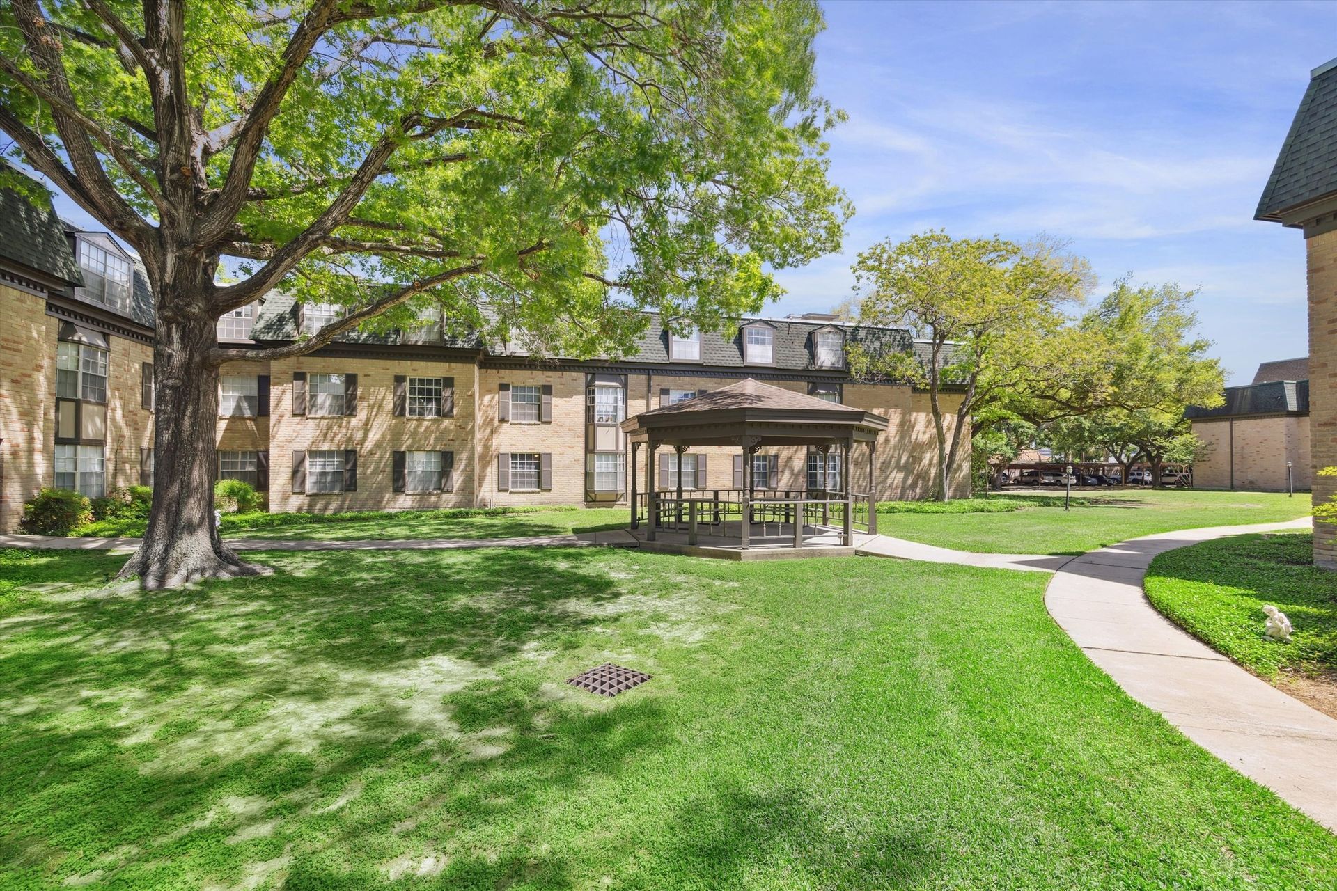 Apartment complex courtyard with gazebo and lush green lawn. Two-story brick buildings under blue sky.