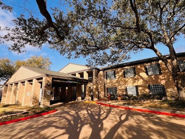 Two-story brick building with a covered entrance, tree canopy overhead, and a red-lined driveway.