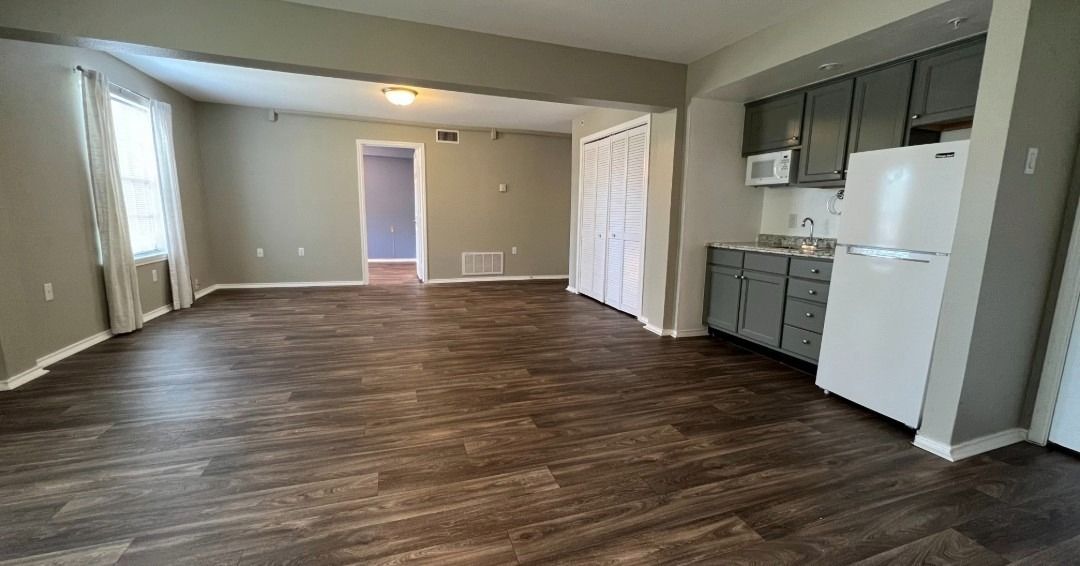 Empty living space with gray walls, dark wood-look floor, and a kitchen area with cabinets and appliances.