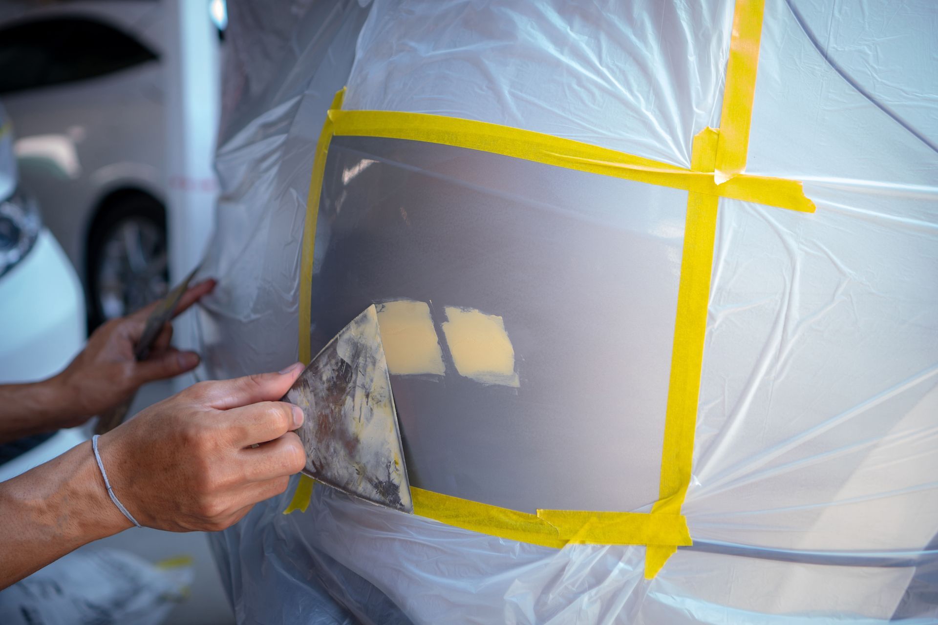 Person applying body filler to a car panel, using a putty knife, within a taped plastic sheet enclosure.