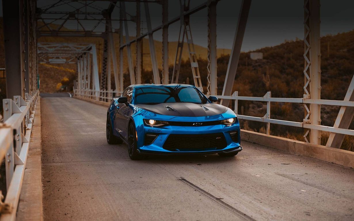 Blue sports car driving on a bridge with metal truss beams and a wooded hillside background