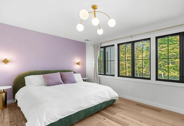 A bedroom with a purple wall , a bed , two windows and a chandelier.