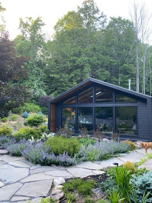 A black house with a lot of windows is surrounded by trees and flowers.
