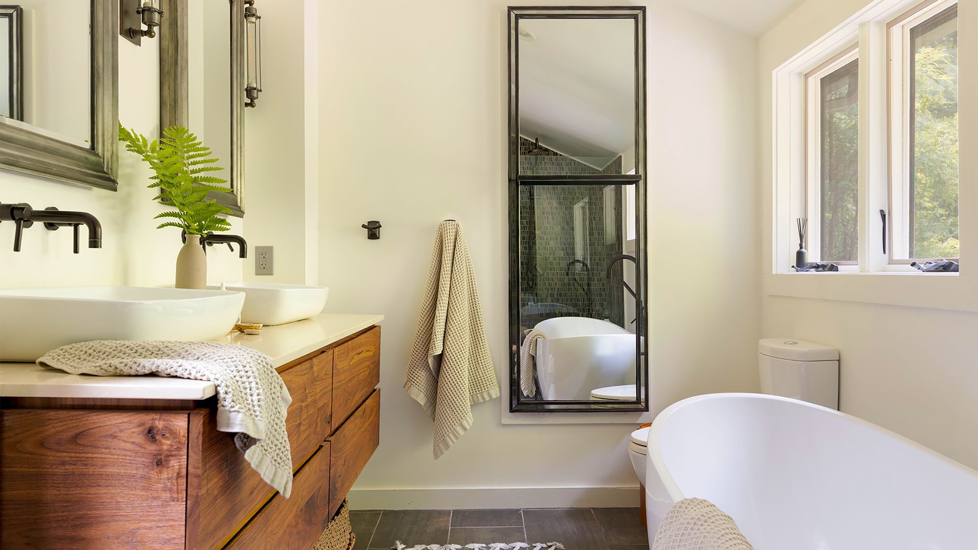 A bright bathroom with a wood vanity, double vessel sinks, a full-length floor mirror, and a standalone soaking tub