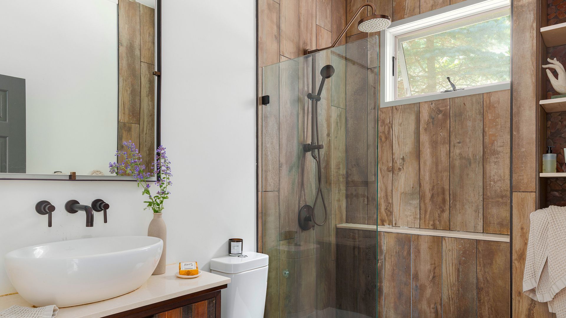 A modern bathroom featuring a white vessel sink on a vanity, a glass shower with wood-paneled walls, and a large mirror