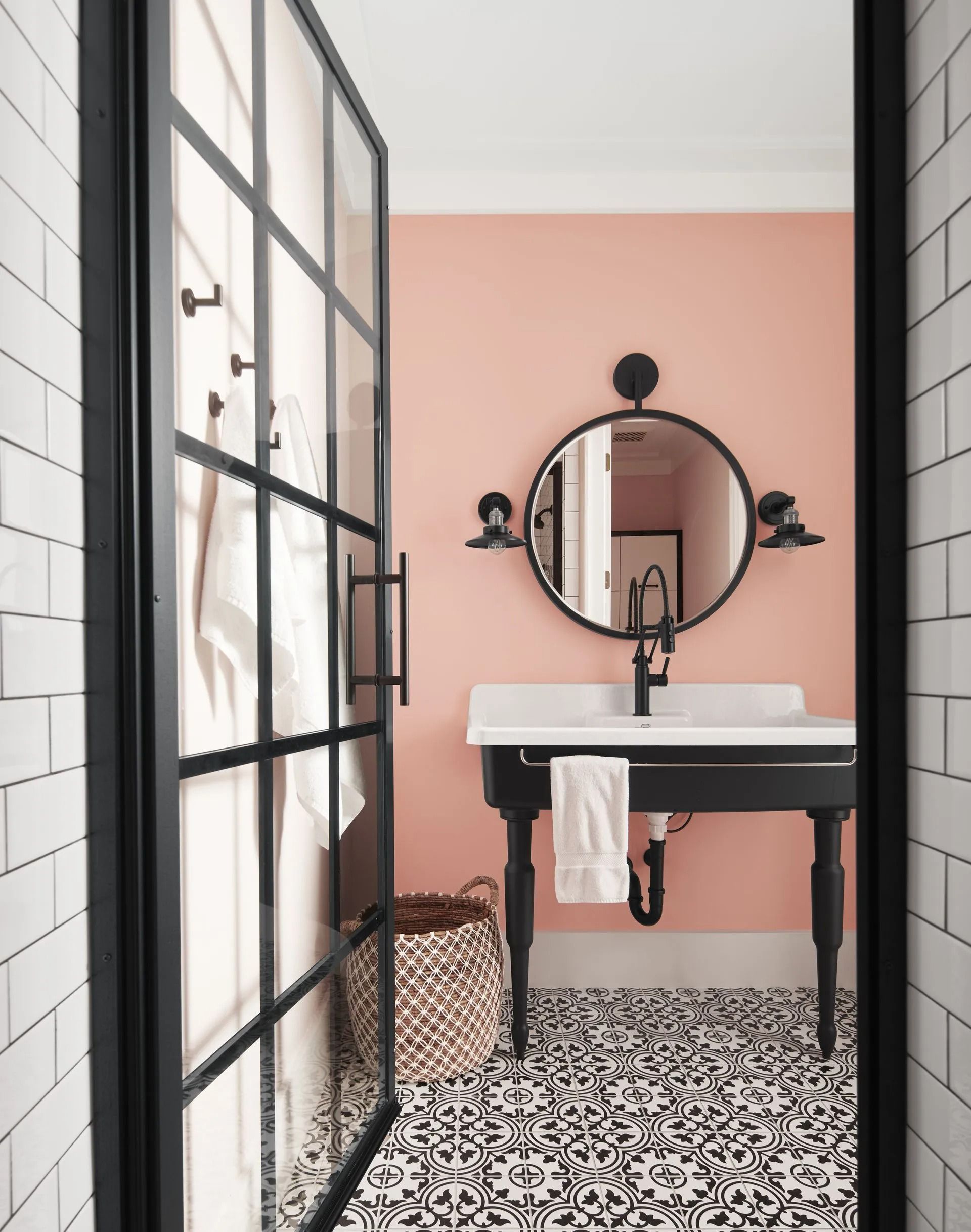 Modern bathroom with pink walls, patterned tile floor, round mirror, and black-framed glass shower door