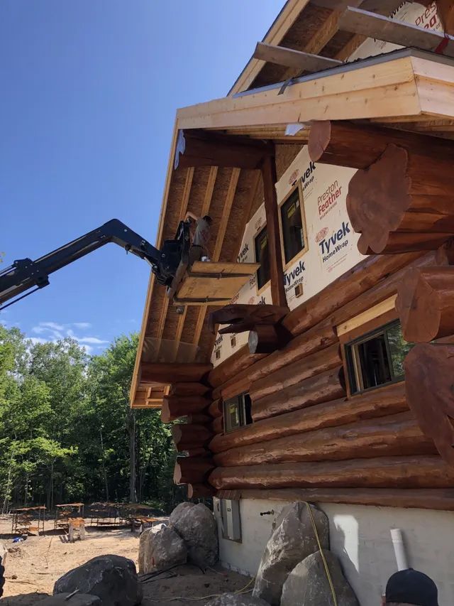 Construction worker on a lift installing roofing material on a log cabin under a blue sky.