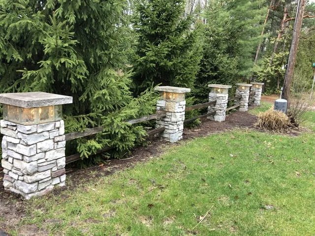 Stone pillars with lights, supporting a wooden fence, in front of evergreen trees and a grassy lawn.