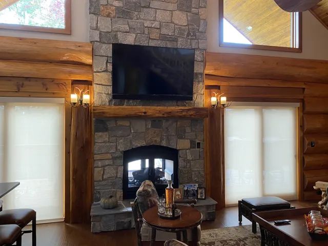 Stone fireplace with TV mounted above, flanked by sconces and windows, in a log cabin style room.