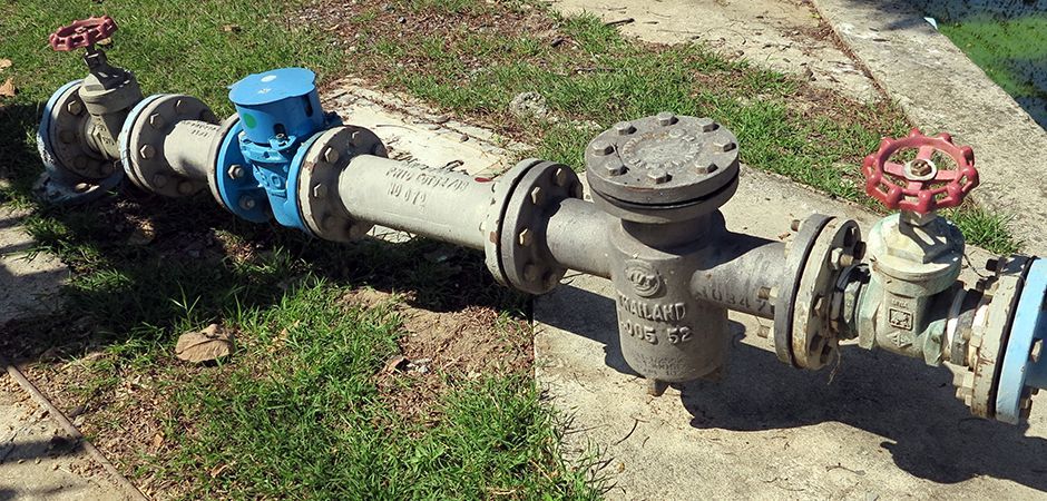 A metal water pipe assembly features a blue meter, a strainer, and two red handwheel valves, situated outdoors on grass.
