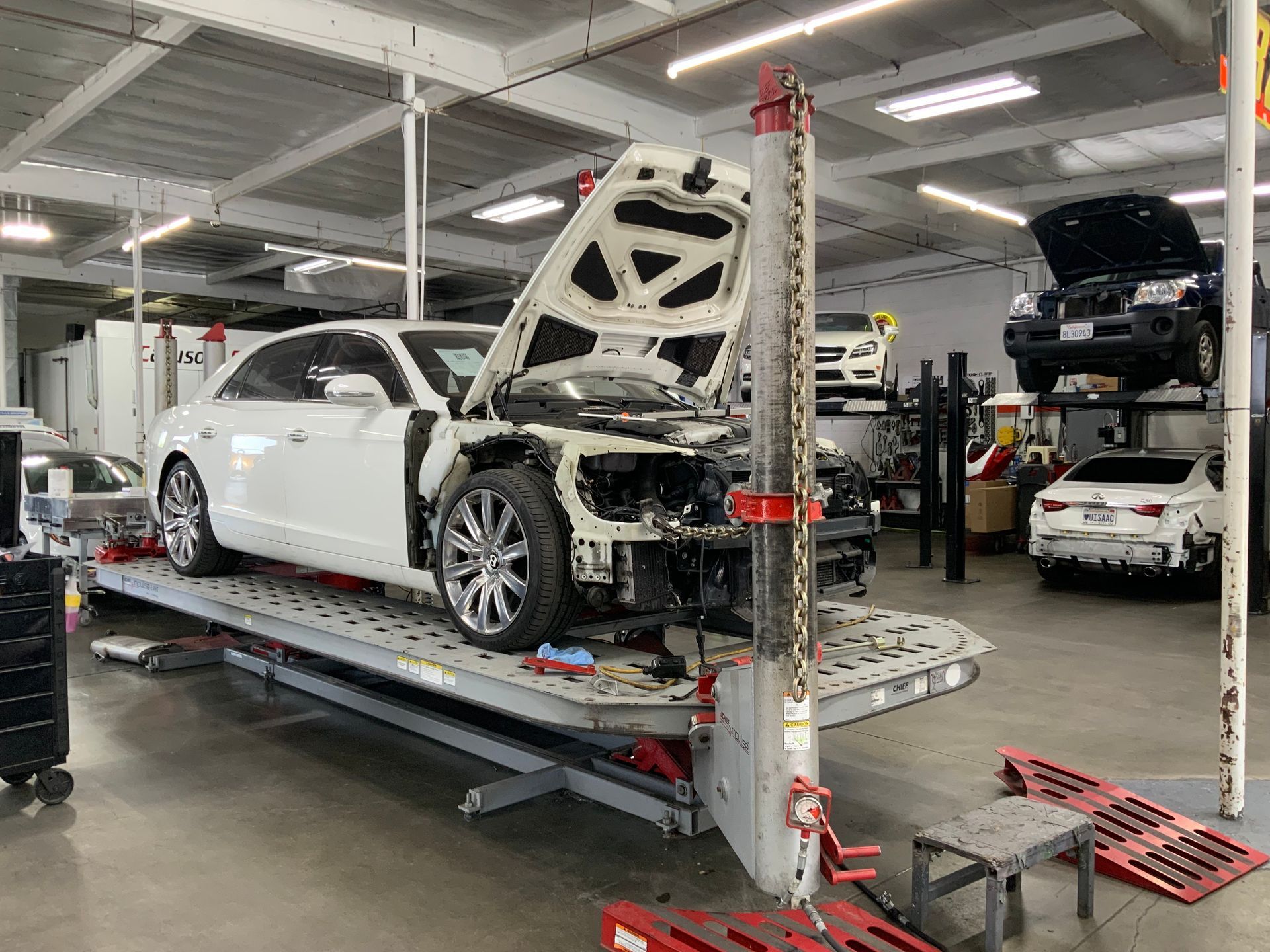 White car undergoing repair in an auto body shop; hood open, parts removed, on a lift.