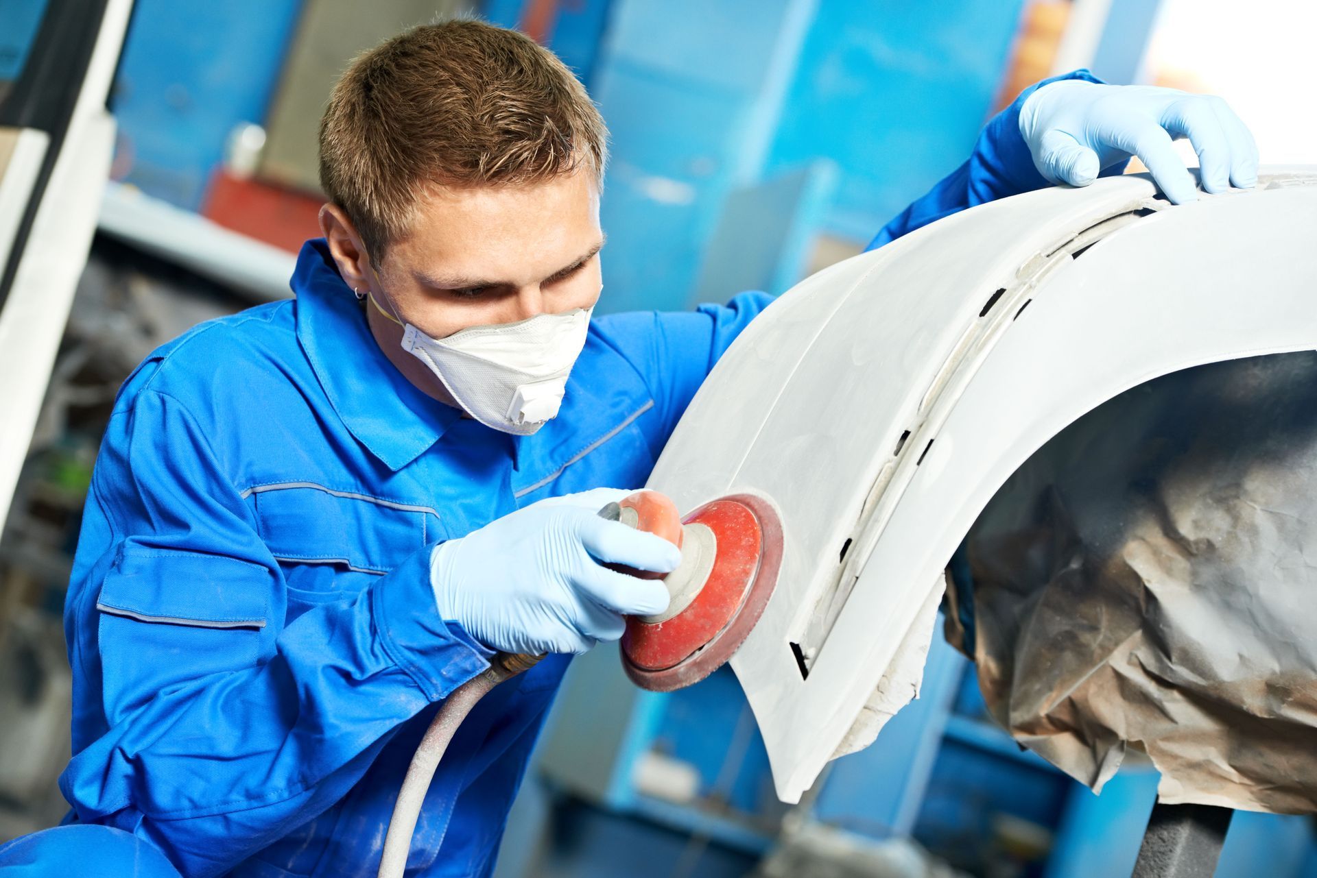 Person in blue jumpsuit sands a car bumper, wearing gloves and a respirator mask.