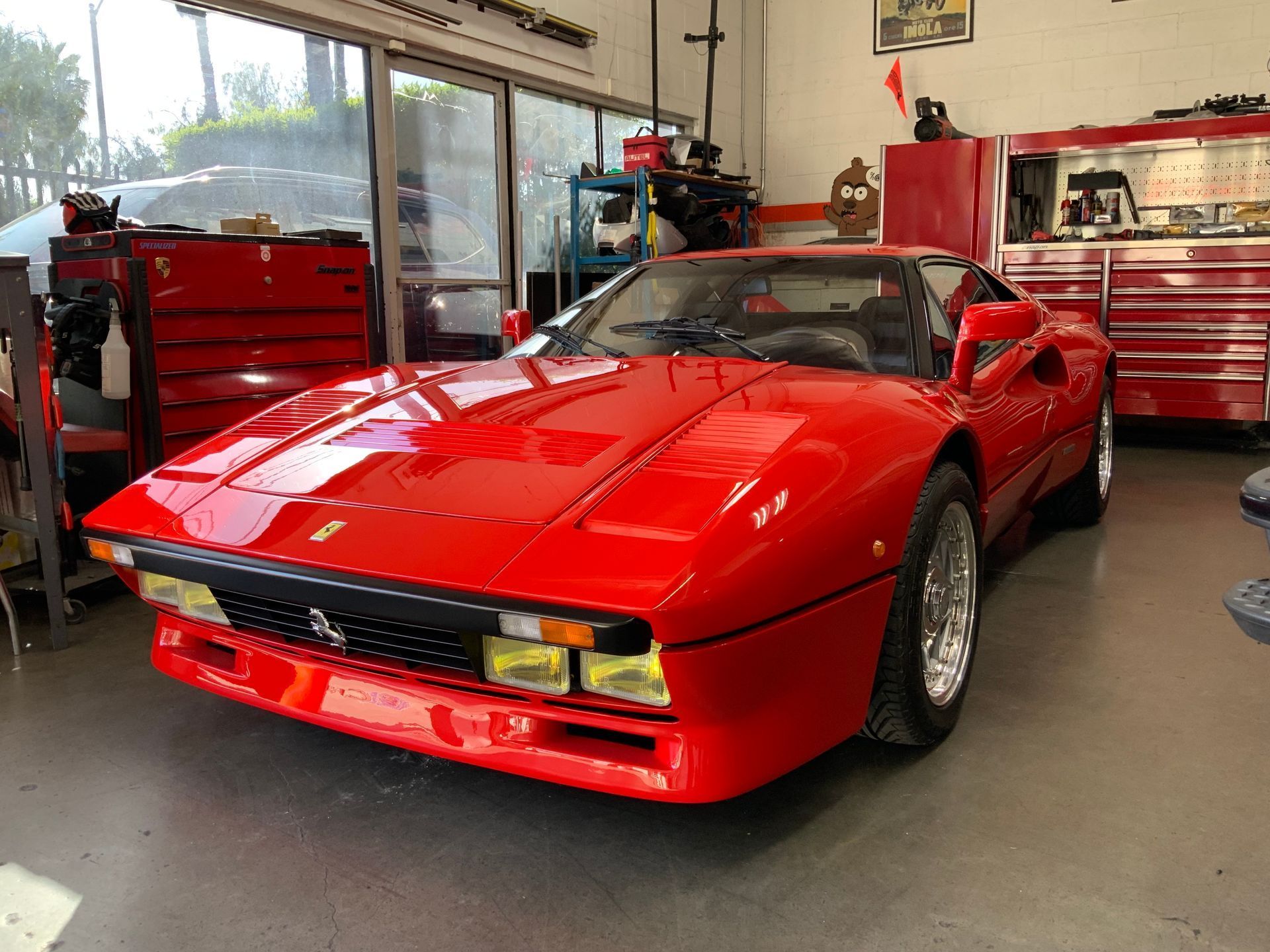 Red sports car parked in a garage, with tool cabinets and windows in the background