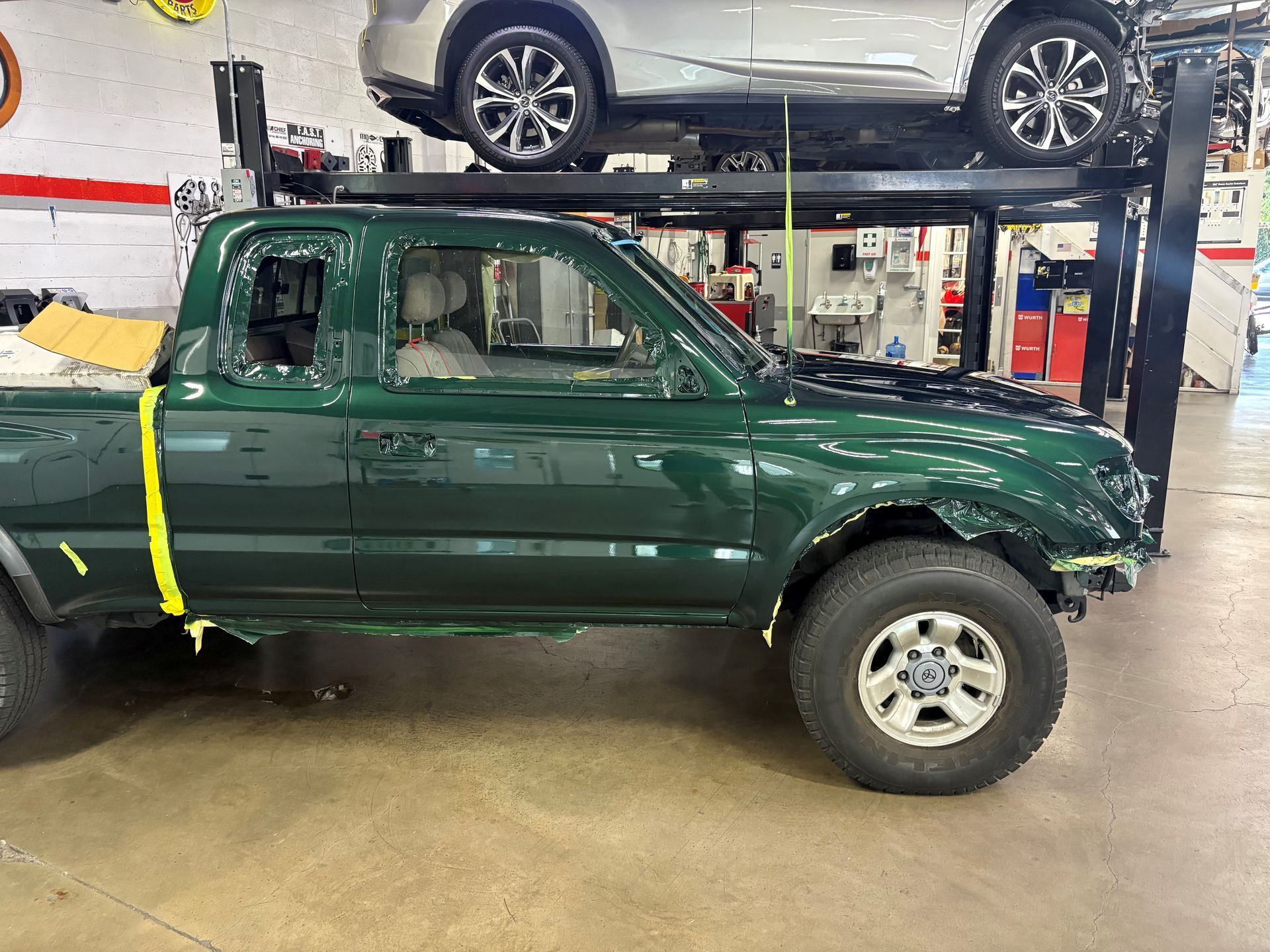 Green pickup truck in an auto shop, strapped down on a lift with a car above it