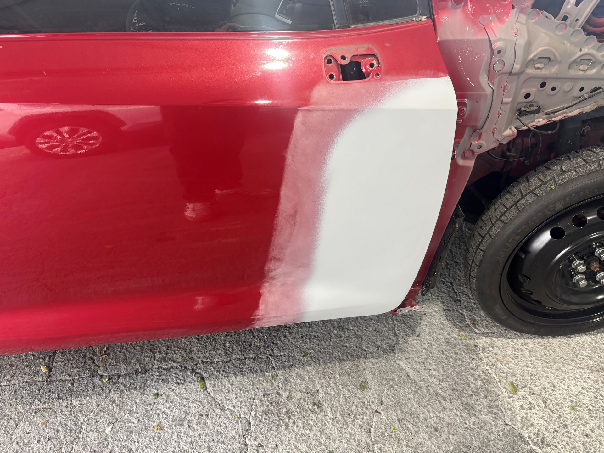 Red car door with primer patch near rear wheel on a concrete driveway