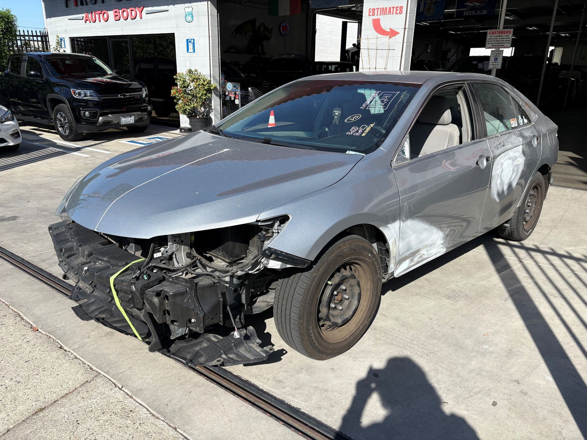 Damaged gray car with front-left bumper and headlight missing, parked in a driveway or lot