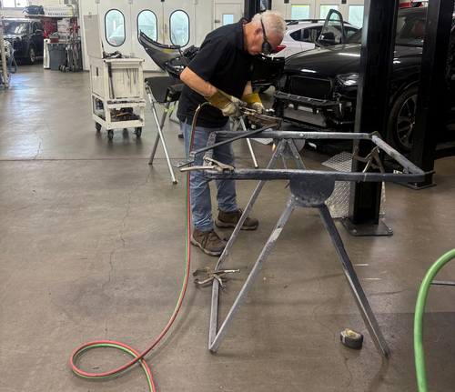A worker sanding a car frame in an auto body shop, with tools and equipment nearby.