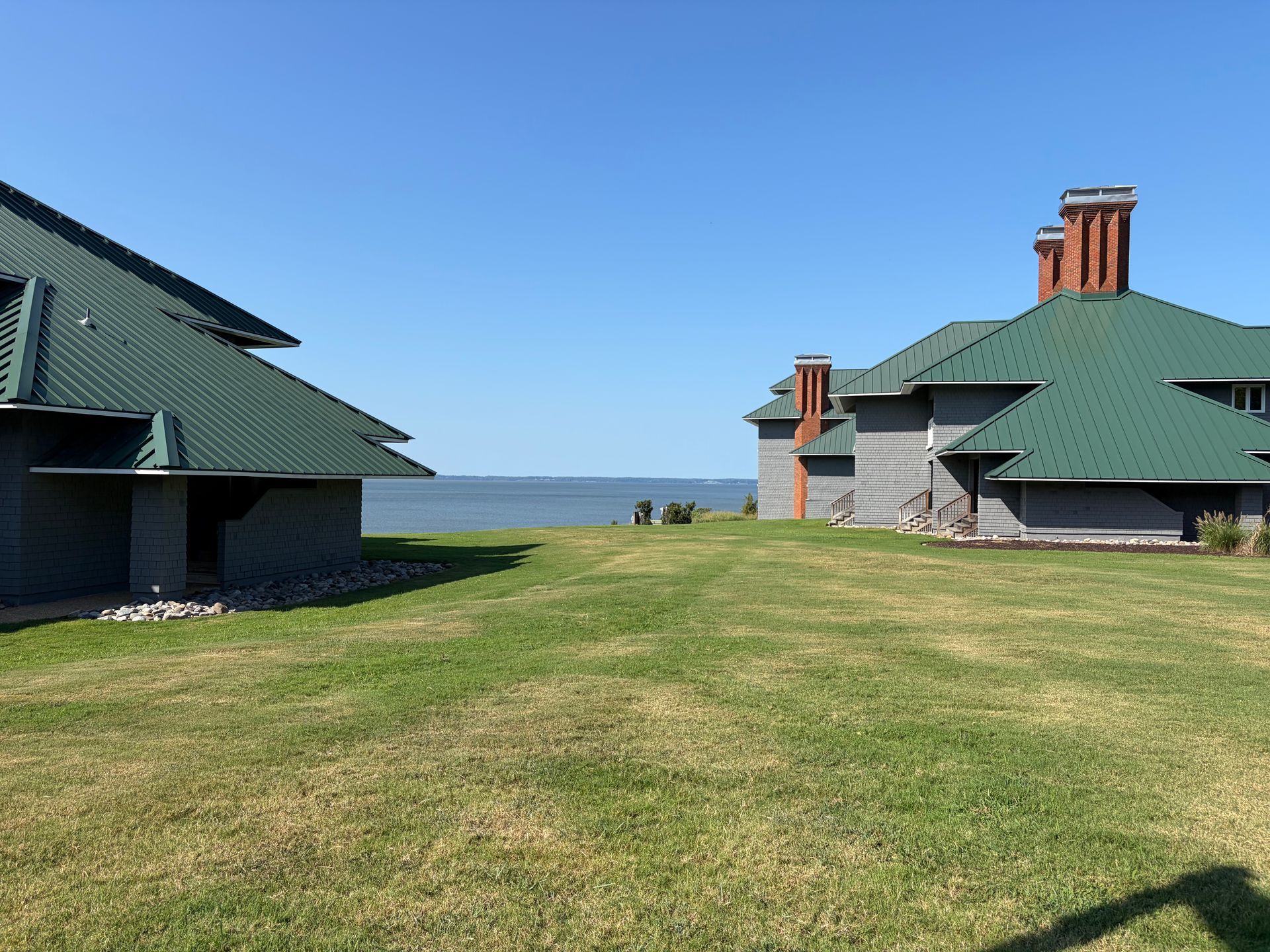 Two green-roofed buildings beside a grassy courtyard overlooking the ocean under a clear blue sky